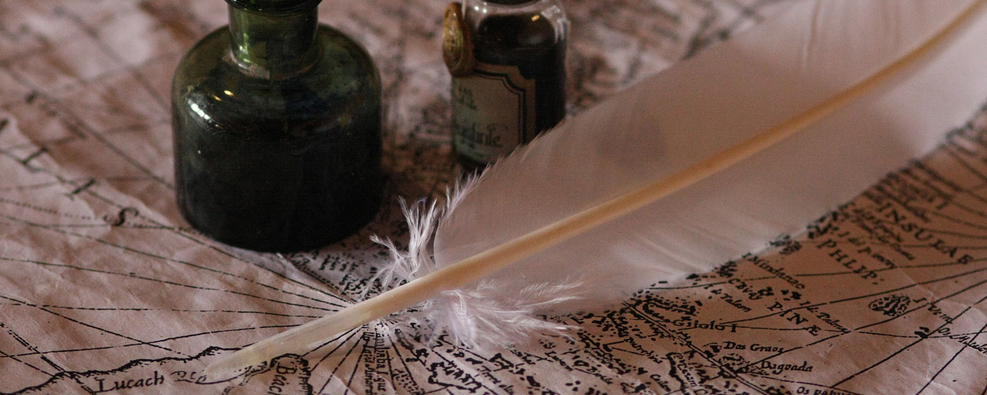 Close-up photograph of a vintage white quill pen resting diagonally on an aged, detailed treasure map, with two glass ink bottles in the background.